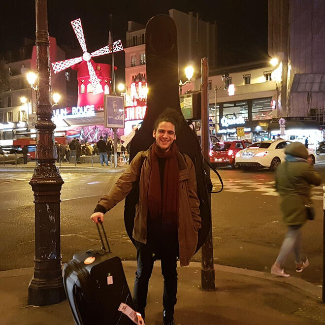 Untitled Elias Kokkoris carrying his double bass and a suitcase after arriving in Paris, with the Moulin Rouge windmill visible in the background.